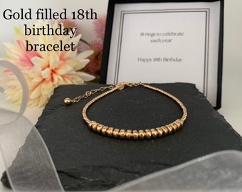 Puede incluir: Un brazalete dorado con 18 cuentas, presentado en una caja de regalo negra. El brazalete está sobre una pizarra gris oscura, con una flor rosa y blanca suave y una cinta gris en el fondo. La caja dice "18 anillos para celebrar cada año. Feliz 18 cumpleaños."