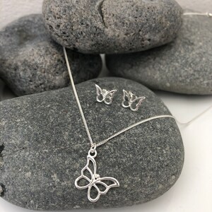 May include: A silver butterfly pendant necklace and matching stud earrings. The jewelry is displayed on a gray stone surface. The butterfly pendant and earrings have an openwork design, showcasing the butterfly shape.