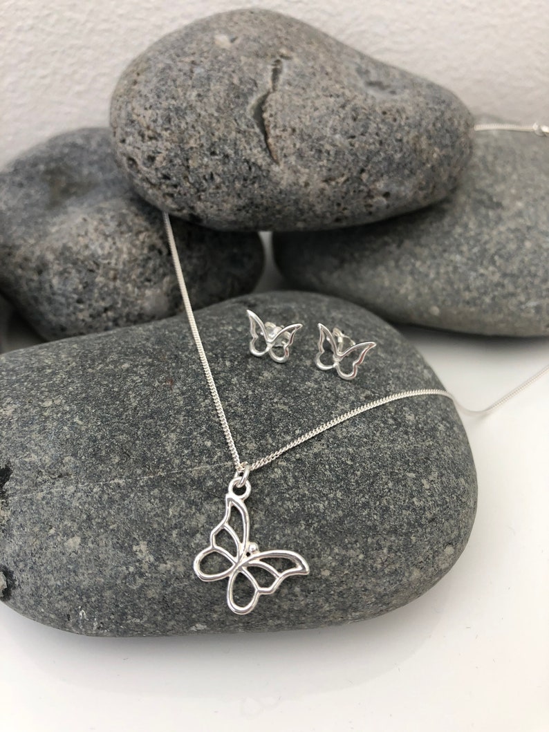 May include: A silver butterfly pendant necklace and matching stud earrings. The jewelry is displayed on a gray stone surface. The butterfly pendant and earrings have an openwork design, showcasing the butterfly shape.