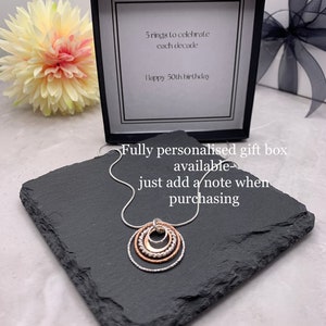 May include: A silver necklace with five interlocking rings, each a different size. The rings are made of silver and rose gold. The necklace is in a black gift box with a white card that says "5 rings to celebrate each decade Happy 50th birthday". The box is on a black slate surface. Fully personalised gift box available just add a note when purchasing.