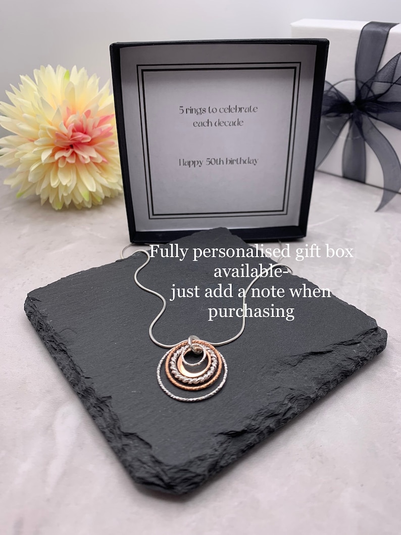 May include: A silver necklace with five interlocking rings, each a different size. The rings are made of silver and rose gold. The necklace is in a black gift box with a white card that says "5 rings to celebrate each decade Happy 50th birthday". The box is on a black slate surface. Fully personalised gift box available just add a note when purchasing.