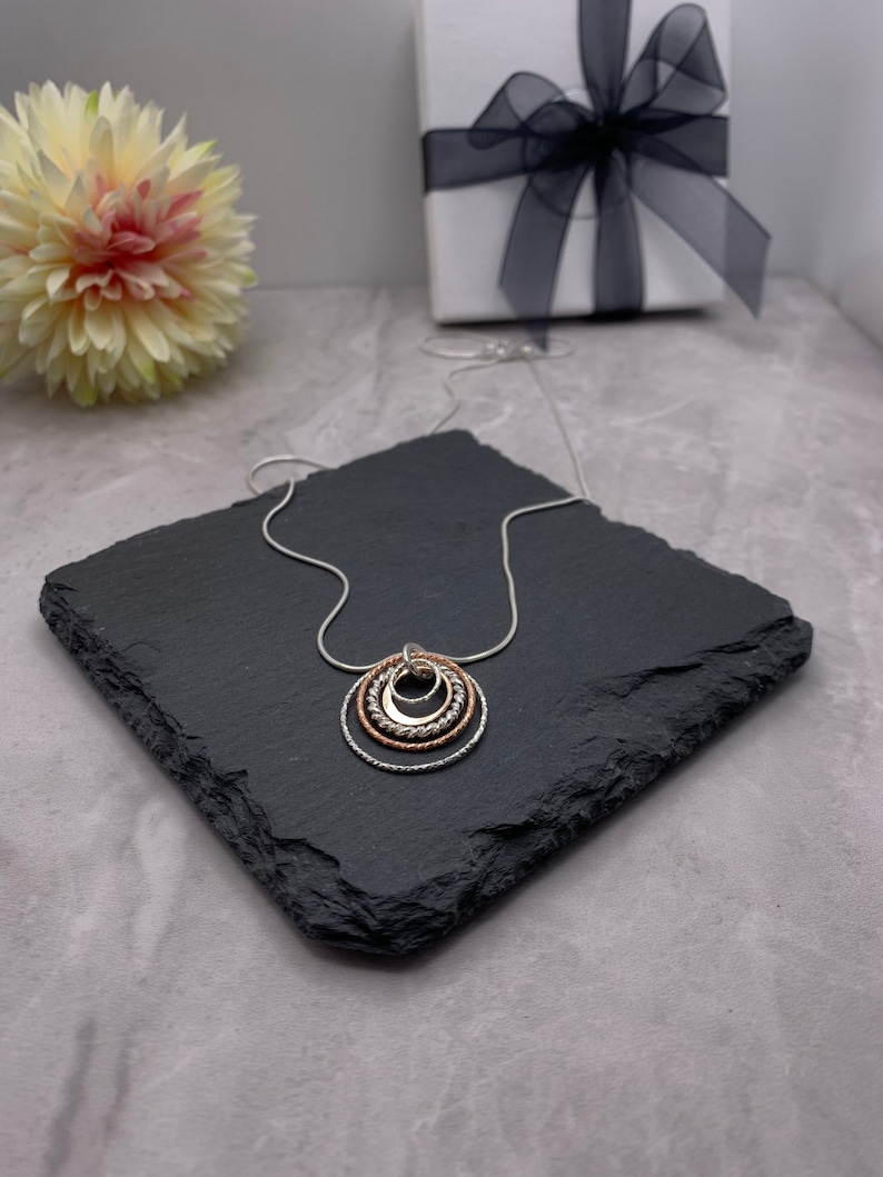May include: A silver chain necklace with a pendant made of three interlocking circles. The circles are silver, rose gold, and silver, and they are textured.