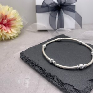 Puede incluir: Un brazalete de plata con una banda lisa y redondeada y m&uacute;ltiples peque&ntilde;as cuentas de plata. El brazalete se muestra sobre una superficie de pizarra negra.