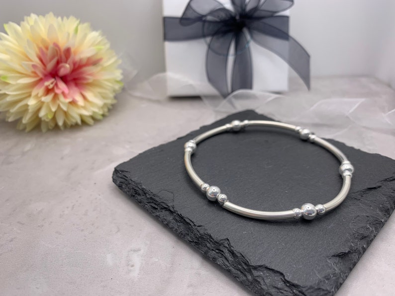 Puede incluir: Un brazalete de plata con una banda lisa y redondeada y m&uacute;ltiples peque&ntilde;as cuentas de plata. El brazalete se muestra sobre una superficie de pizarra negra.