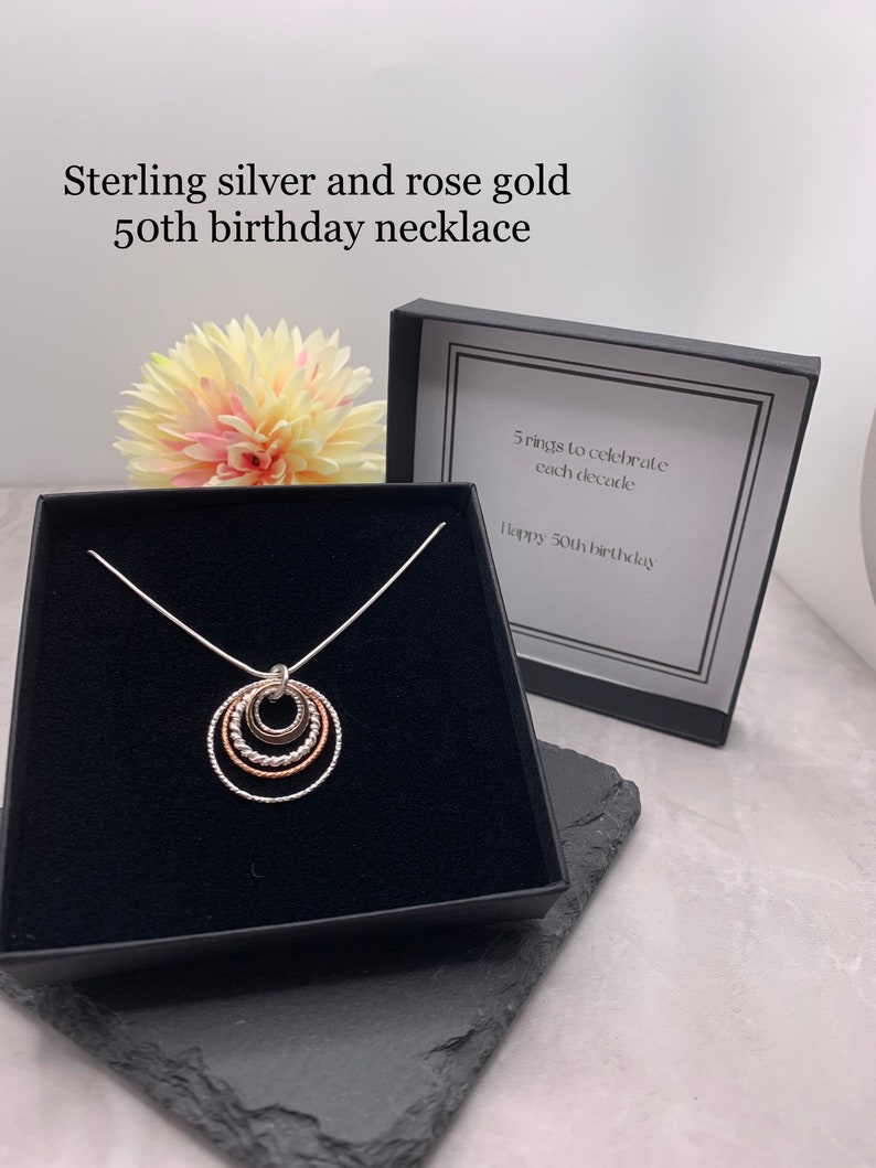 May include: Sterling silver and rose gold necklace with five interlocking rings, representing each decade of a 50th birthday. The necklace is displayed in a black box with a white card that reads "5 rings to celebrate each decade. Happy 50th birthday."