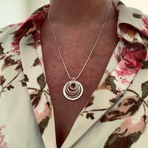 Pu&ograve; includere: Una collana d'argento con un ciondolo composto da cinque cerchi intrecciati. La collana &egrave; indossata da una persona che indossa una blusa floreale.