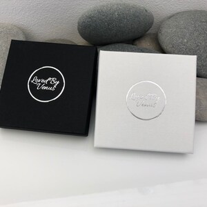 May include: Two square boxes with the text "Loved By Venus" in a circle on the top of each box. One box is black with silver text and the other box is white with silver text.