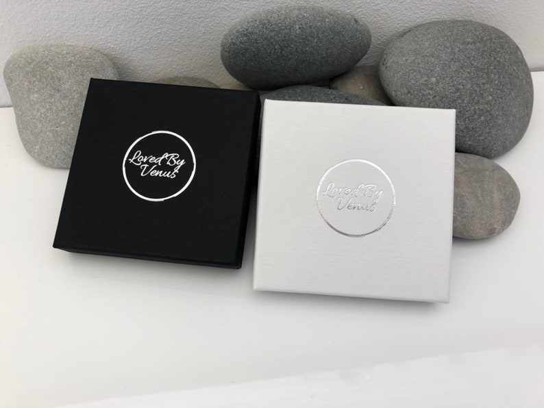 May include: Two square boxes with the text "Loved By Venus" in a circle on the top of each box. One box is black with silver text and the other box is white with silver text.