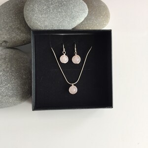 May include: A set of rose quartz earrings and a necklace in a black box. The earrings and necklace feature a round, light pink rose quartz stone.