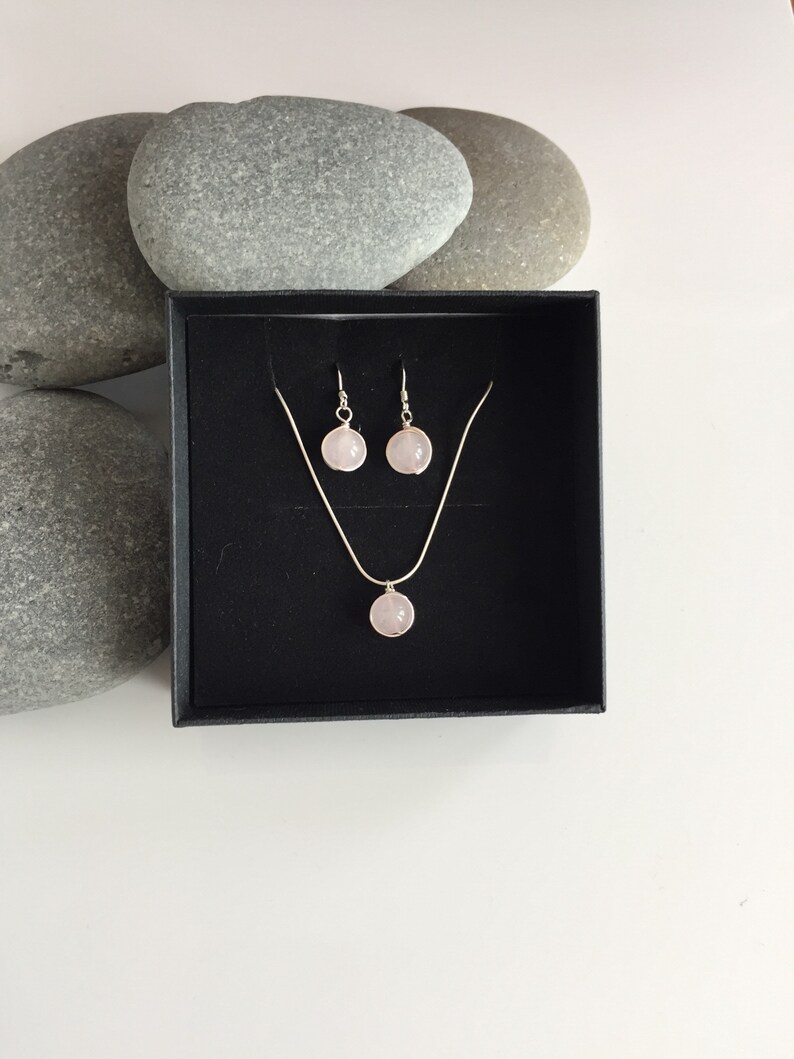May include: A set of rose quartz earrings and a necklace in a black box. The earrings and necklace feature a round, light pink rose quartz stone.