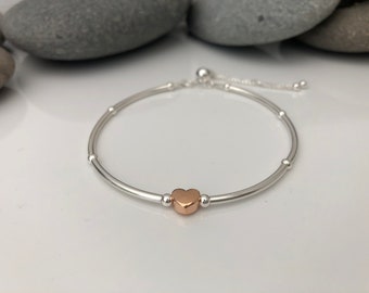 Puede incluir: Una pulsera de plata con un dije de corazón de oro rosa y pequeñas cuentas de plata. La pulsera es ajustable con un cierre deslizante.