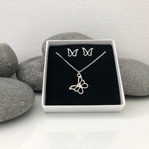 May include: A silver butterfly jewelry set, including a necklace and matching earrings, presented in a white box with a black velvet interior. The butterfly pendant hangs from a delicate chain. The earrings are small butterfly studs.