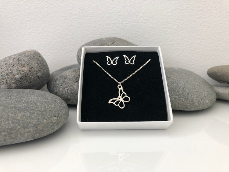 May include: A silver butterfly jewelry set, including a necklace and matching earrings, presented in a white box with a black velvet interior. The butterfly pendant hangs from a delicate chain. The earrings are small butterfly studs.