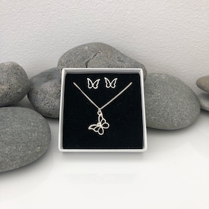 May include: A silver-colored butterfly jewelry set, including a necklace and matching earrings, presented in a white box with a black interior. The butterfly pendant and earrings have an openwork design. The set is displayed against a backdrop of gray stones.
