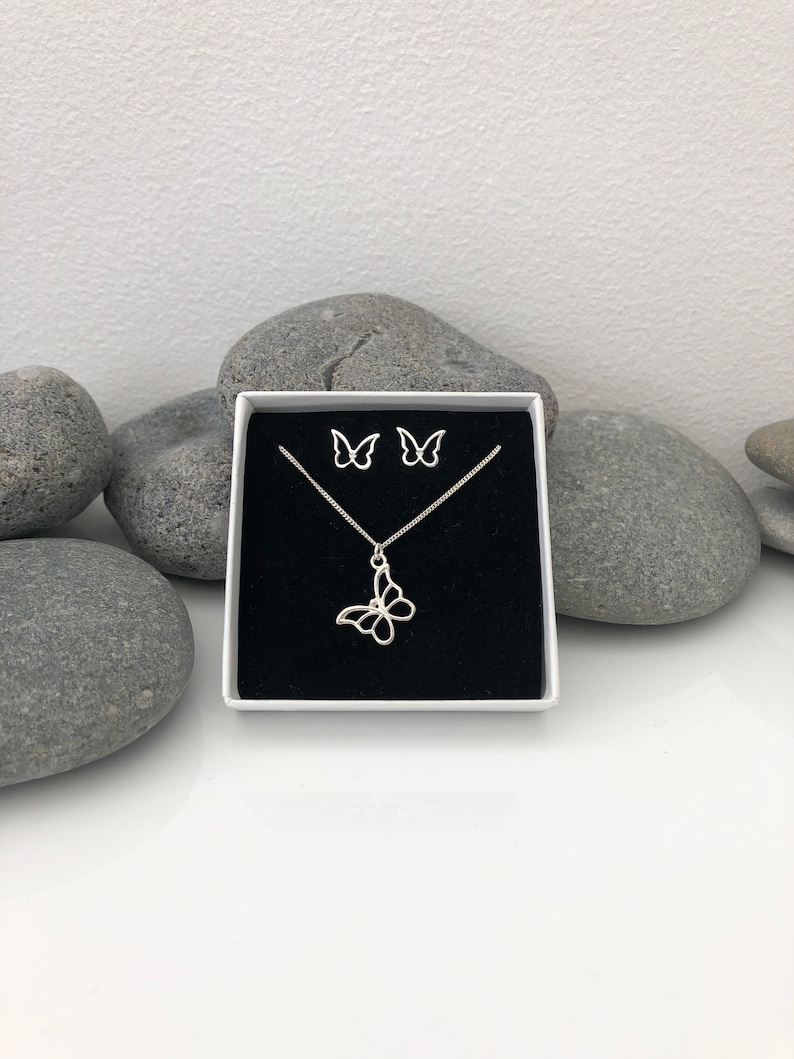 May include: A silver-colored butterfly jewelry set, including a necklace and matching earrings, presented in a white box with a black interior. The butterfly pendant and earrings have an openwork design. The set is displayed against a backdrop of gray stones.
