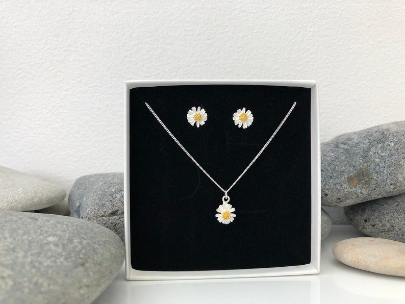 K&ouml;nnte beinhalten: Eine wei&szlig;e Schachtel mit schwarzem Innenraum zeigt eine silberne Halskette und Ohrringe mit einem wei&szlig;en G&auml;nsebl&uuml;mchen-Design. Die Halskette hat einen einzelnen G&auml;nsebl&uuml;mchen-Anh&auml;nger und die Ohrringe haben jeweils ein einzelnes G&auml;nsebl&uuml;mchen.