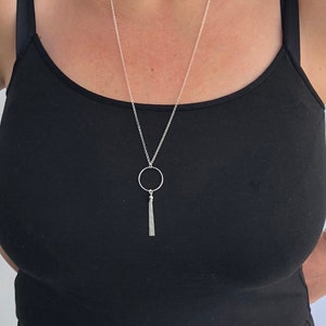 May include: A silver chain necklace with a circle pendant and a tassel. The necklace is on a person wearing a black tank top.