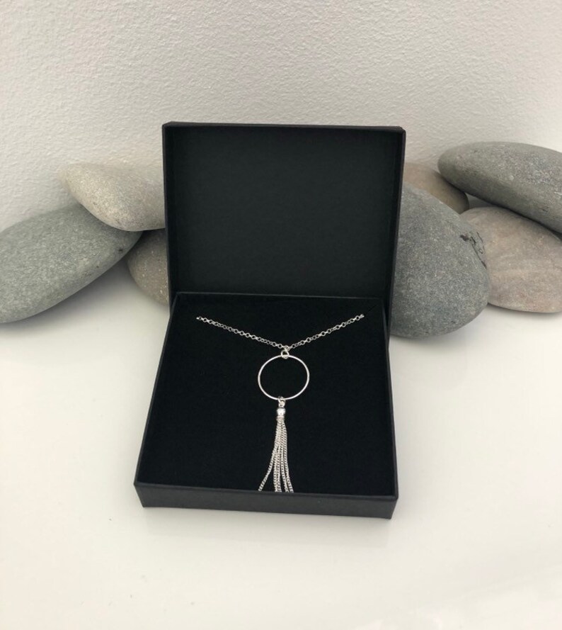 May include: A silver necklace with a circle pendant and a tassel. The necklace is in a black box.
