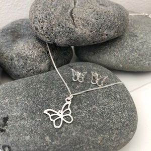 May include: A silver butterfly pendant necklace and matching stud earrings. The jewelry is displayed on a gray stone surface. The butterfly pendant and earrings have an openwork design. The necklace chain is delicate and silver.