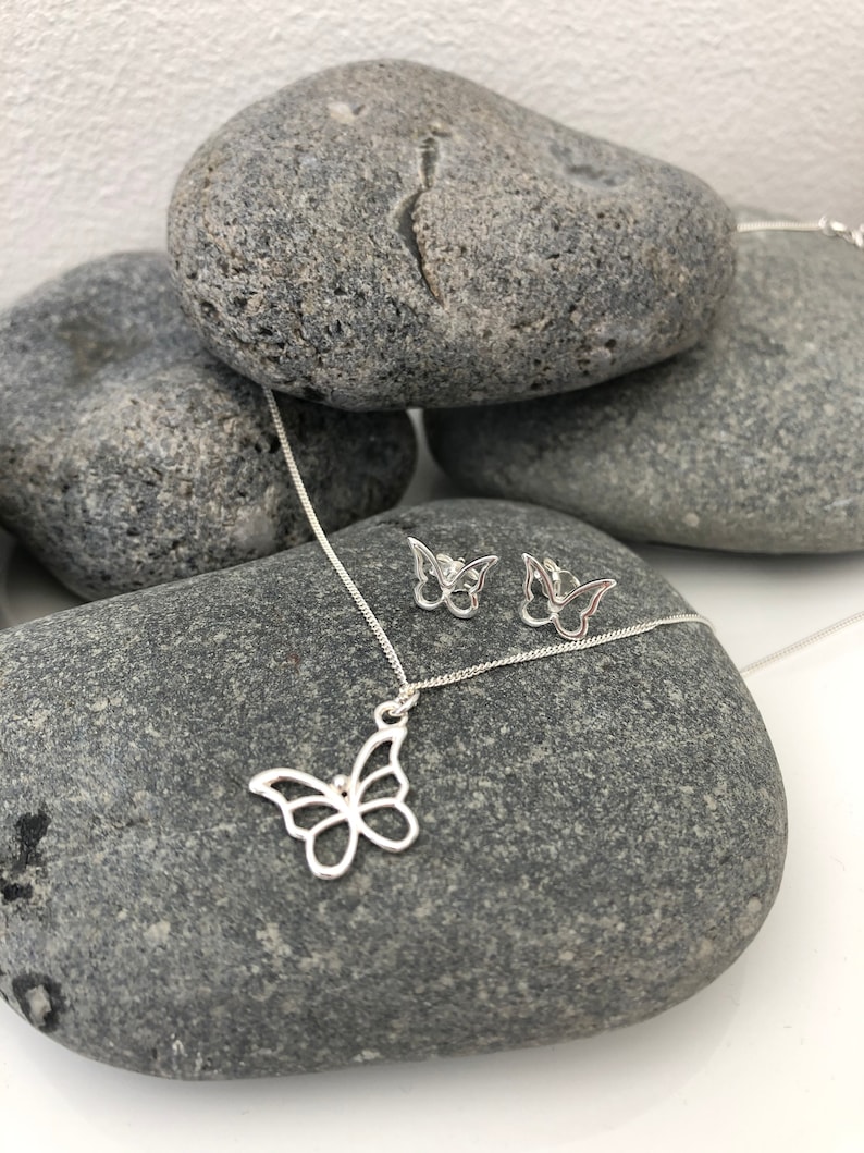 May include: A silver butterfly pendant necklace and matching stud earrings. The jewelry is displayed on a gray stone surface. The butterfly pendant and earrings have an openwork design. The necklace chain is delicate and silver.