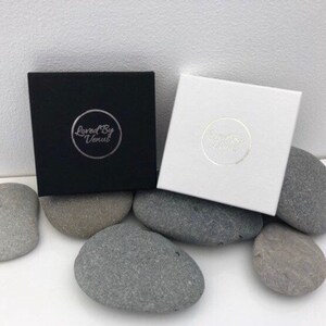 May include: Two square boxes, one black and one white, each with a circular logo, rest on gray stones. The black box has the text "Loved By Venus" in a silver circle. The white box has a similar logo. The boxes are on a white surface.