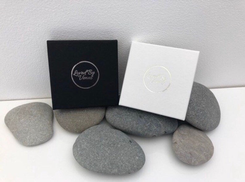 May include: Two square boxes, one black and one white, each with a circular logo, rest on gray stones. The black box has the text "Loved By Venus" in a silver circle. The white box has a similar logo. The boxes are on a white surface.