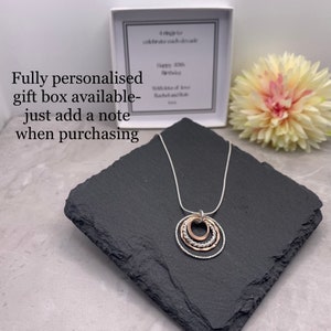 May include: A silver chain necklace with a pendant made of three interlocking circles. The outer two circles are silver, and the inner circle is rose gold. The necklace is displayed on a black slate surface.