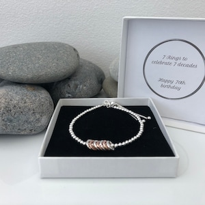 May include: A silver bracelet with seven rose gold rings, presented in a white box with the text "7 rings to celebrate 7 decades Happy 70th birthday".