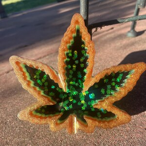 Op de afbeelding: Een decoratief harsblad in de vorm van een cannabisblad. Het blad heeft een gouden glitterrand, een groen en oranje interieur en een donker midden gevuld met groene stervormige glitter.