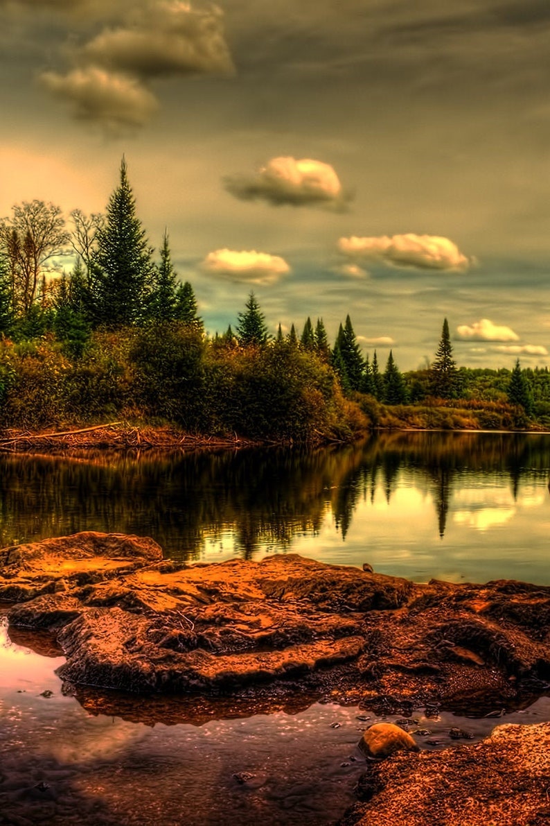 May include: A scenic landscape with a still lake reflecting the cloudy sky and a line of evergreen trees along the shore. A large, brown rock sits in the foreground.