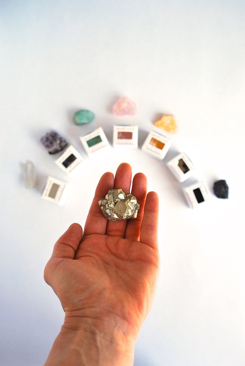 May include: A hand holding a gold-colored pyrite crystal. The crystal is surrounded by small boxes containing various colored crystals.