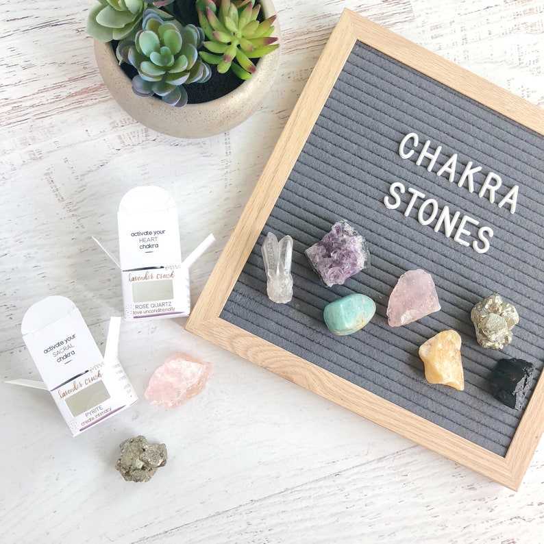 K&ouml;nnte beinhalten: Ein Set Chakra-Steine mit einem Buchstabenbrett, auf dem "Chakra-Steine" steht. Die Steine sind Amethyst, Rosenquarz, T&uuml;rkis, Citrin, Pyrit und schwarzer Obsidian. Die Steine sind in einer Reihe auf dem Buchstabenbrett angeordnet. Es gibt zwei kleine Schachteln mit dem Text "Aktiviere dein Herzchakra" und "Aktiviere dein Sakralchakra" darauf.