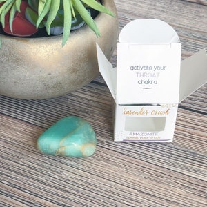 May include: A small, teal green amazonite crystal in a white box with a clear window. The box has the text "activate your throat chakra" and "lavender crush" on it. The crystal is labeled "amazonite speak your truth".