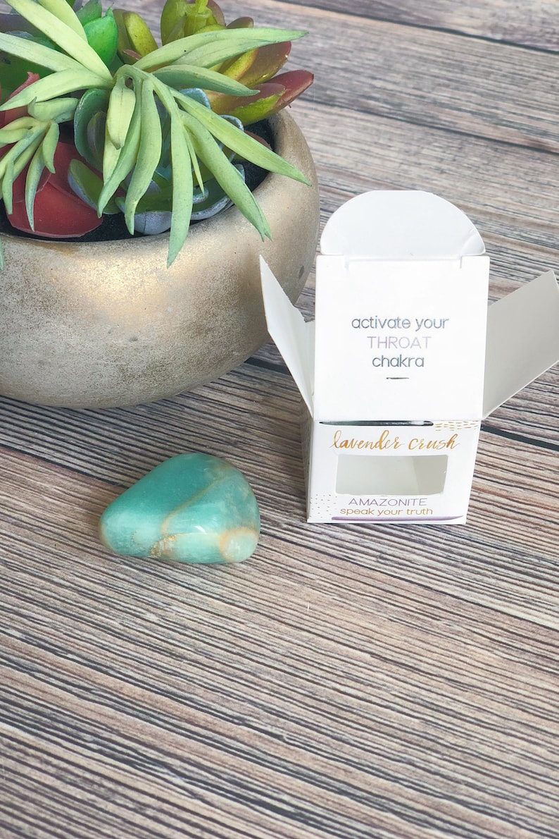 May include: A small, teal green amazonite crystal in a white box with a clear window. The box has the text "activate your throat chakra" and "lavender crush" on it. The crystal is labeled "amazonite speak your truth".