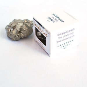 May include: A white box with the text "activate your SACRAL chakra" and "Lavender crush" on it. The box also has the text "live pleasurably, live creatively, live meaningfully" and "LAVENDER CRUSH". A small, gray, metallic rock is next to the box.