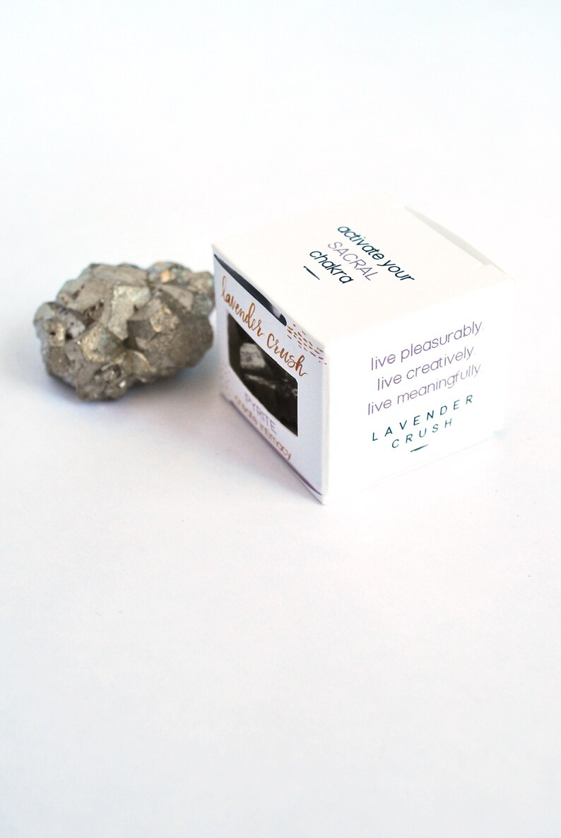 May include: A white box with the text "activate your SACRAL chakra" and "Lavender crush" on it. The box also has the text "live pleasurably, live creatively, live meaningfully" and "LAVENDER CRUSH". A small, gray, metallic rock is next to the box.