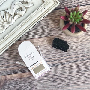 May include: A small white box with the text "activate your ROOT chakra" and "lavender crush" on it. The box contains a small piece of black tourmaline crystal. The text on the box also says "BLACK TOURMALINE stay grounded".