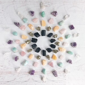 K&ouml;nnte beinhalten: Eine Vielzahl von Kristallen und Steinen, die in einem Kreis auf einer wei&szlig;en Holzoberfl&auml;che angeordnet sind. Die Kristalle umfassen Amethyst, Rosenquarz, klarer Quarz, Pyrit und schwarzer Turmalin.