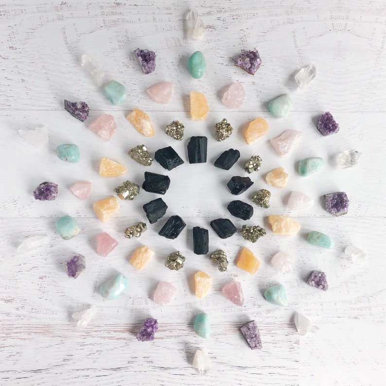 K&ouml;nnte beinhalten: Eine Vielzahl von Kristallen und Steinen, die in einem Kreis auf einer wei&szlig;en Holzoberfl&auml;che angeordnet sind. Die Kristalle umfassen Amethyst, Rosenquarz, klarer Quarz, Pyrit und schwarzer Turmalin.