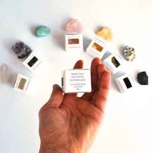 May include: A small white box with the text "speak freely, live truthfully, live meaningfully" and "LAVENDER CRUSH" printed on it. The box is being held in a hand against a white background.