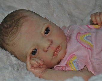 May include: A realistic baby doll with brown hair, blue eyes, and a pink and rainbow patterned outfit.