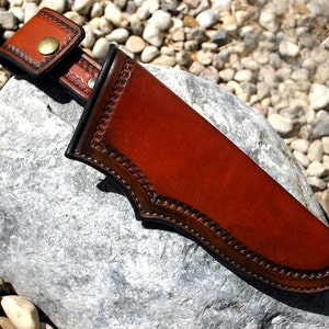 May include: A brown leather knife sheath with a decorative stitched border and a belt loop. The sheath is resting on a gray rock.