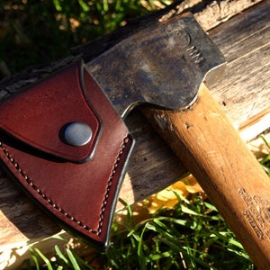 Può includere: Un'ascia vintage con manico in legno e testa in metallo, parzialmente coperta da un fodero in pelle marrone. L'ascia è appoggiata su un pezzo di legno, con erba verde sullo sfondo. La testa dell'ascia ha la scritta "MM".