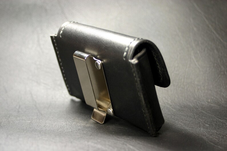 May include: A black leather wallet with a silver metal clip. The wallet has a stitched edge and a flap that closes with a snap.