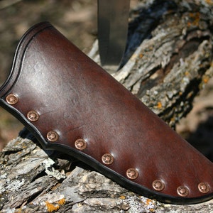 May include: A brown leather knife sheath with copper rivets. The sheath is lying on a piece of bark.