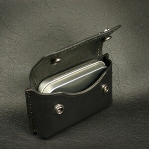May include: A black leather case with a silver metal tin inside. The case has a flap that closes with a snap.