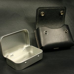 May include: A silver metal tin with a hinged lid is open and next to a black leather pouch with a flap and snap closure. Both items are on a black textured surface.