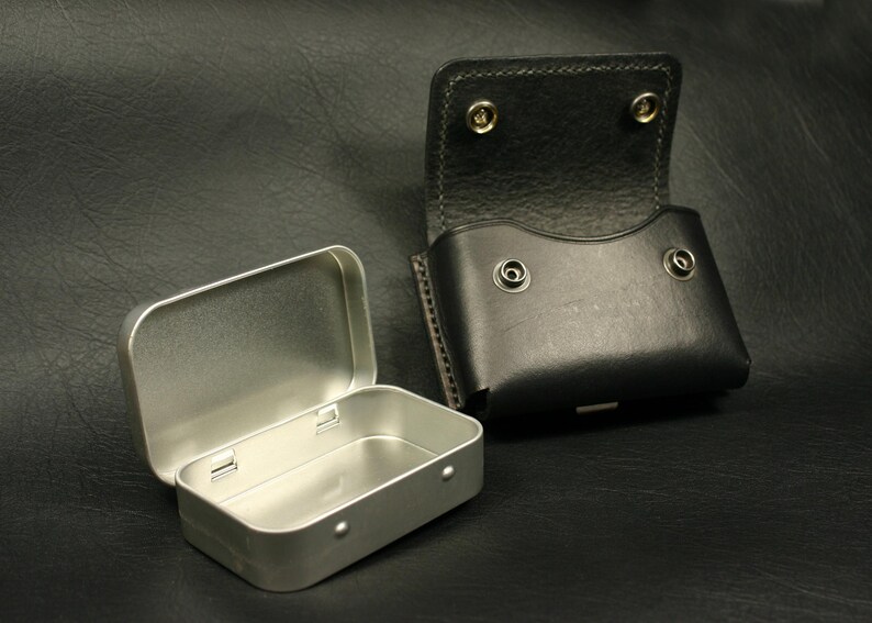 May include: A silver metal tin with a hinged lid is open and next to a black leather pouch with a flap and snap closure. Both items are on a black textured surface.