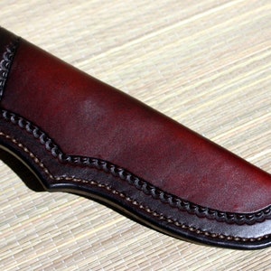 Può includere: Un fodero per coltello in pelle rosso scuro con un bordo decorativo cucito.