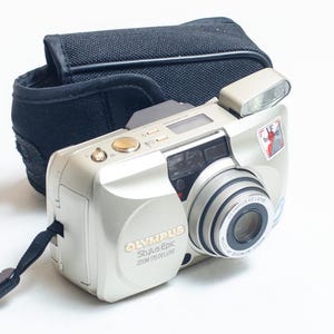 May include: A silver Olympus Stylus Epic Zoom 170 Deluxe film camera with a retractable lens, a built-in flash, and a black wrist strap. The camera is positioned in front of a black fabric carrying case with a flap closure and a textured surface.